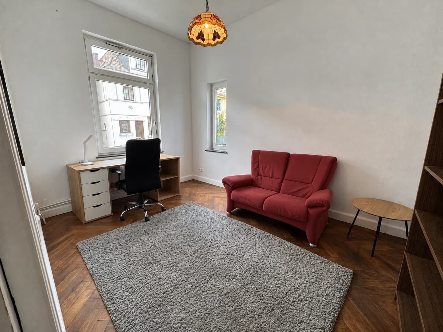 Arbeitszimmer mit Schreibtisch am Fenster, Bürostuhl und rotem Sofa.