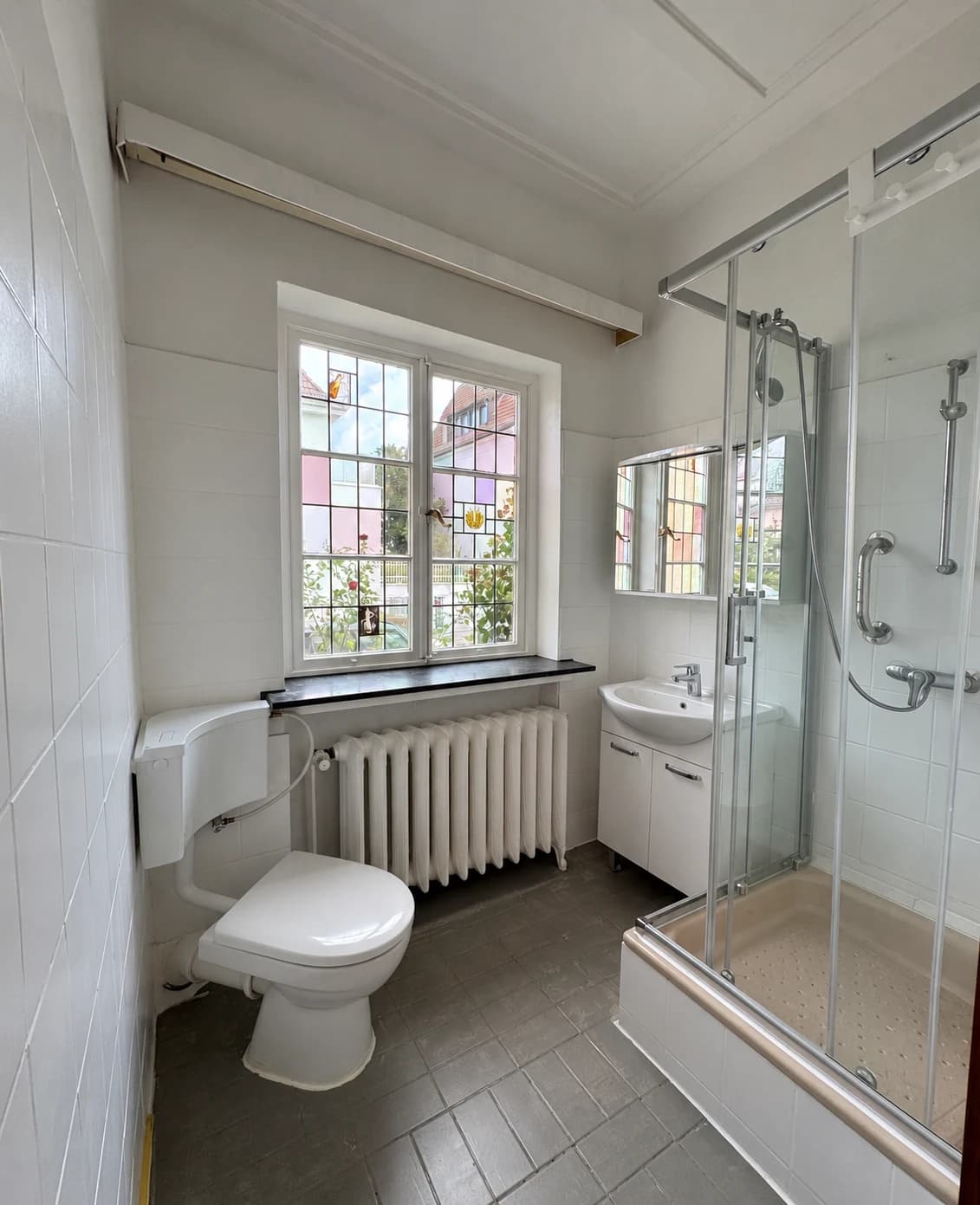 Badezimmer mit Glasdusche, Waschbecken, WC und Fenster.