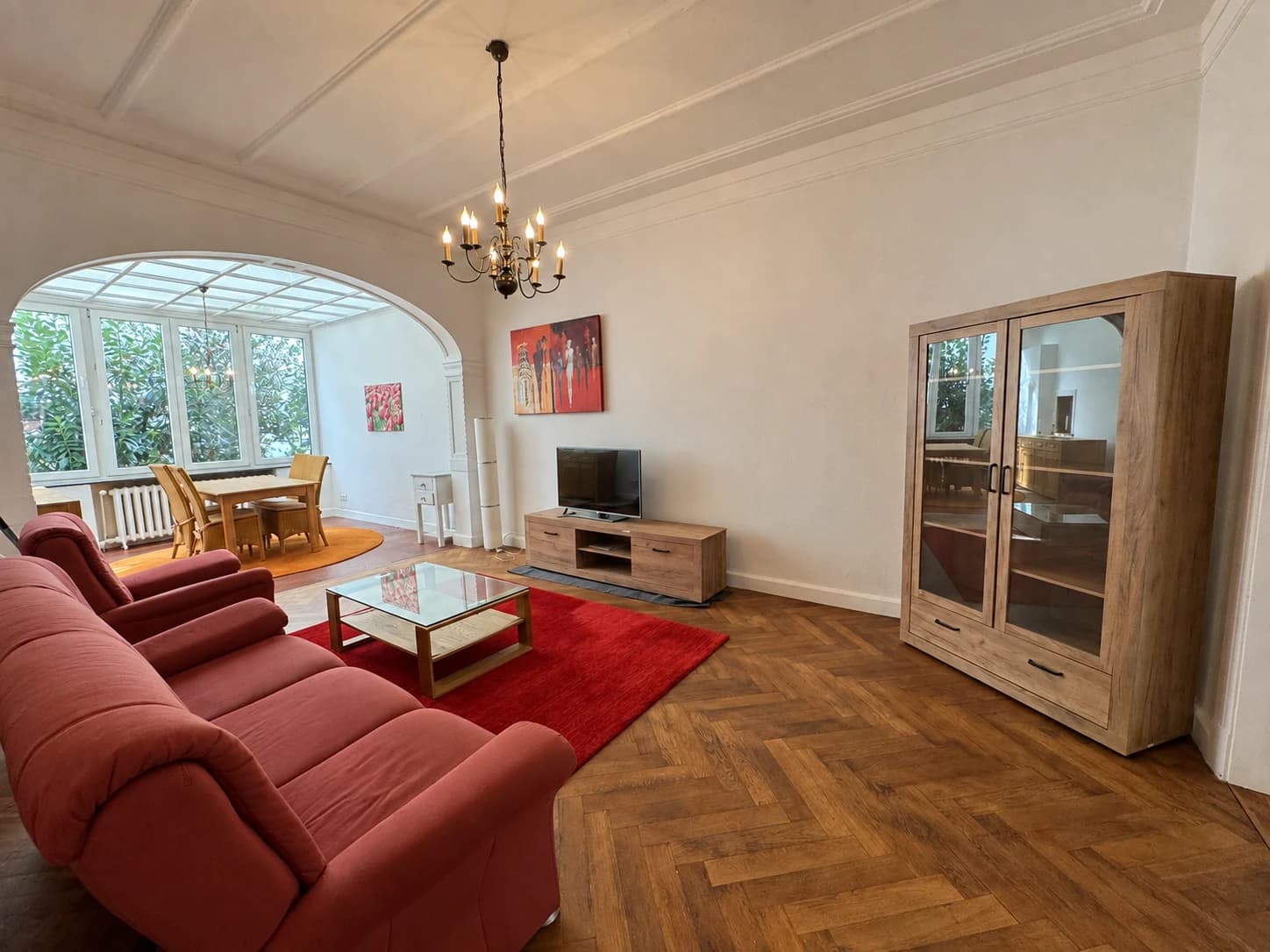 Wohnzimmer mit Sitzgruppe, rotem Teppich, Fernseher und angrenzendem Wintergarten.