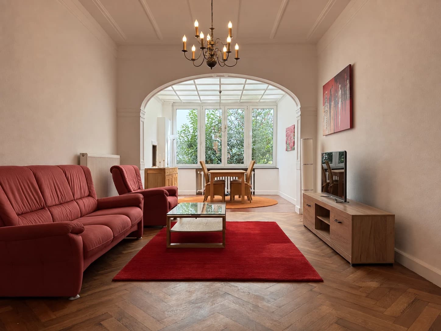 Wohnzimmer mit rotem Sofa, Glastisch und Rundbogen zum Wintergarten.
